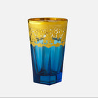 A blue and gold decorative drinking glass with a faceted geometric design, featuring a wide gold band with an intricate cutout pattern around the upper half, set against a plain light gray background.